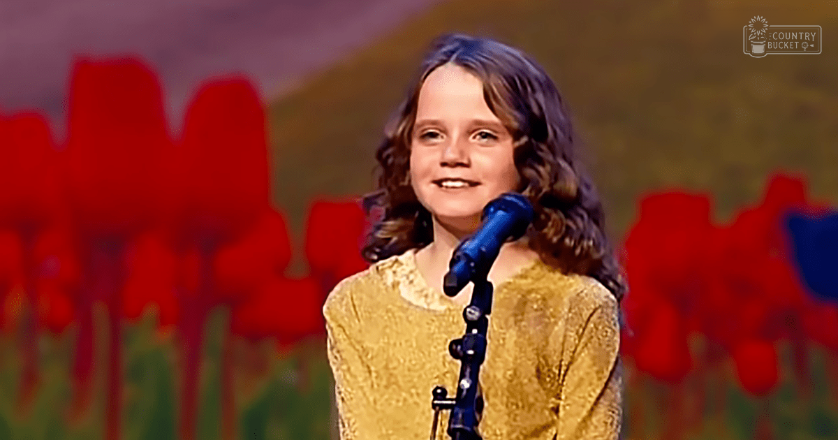 A young adult female with shoulder-length curly hair stands on an indoor stage or performance area wearing a yellow, textured long-sleeved garment under artificial evening lighting.