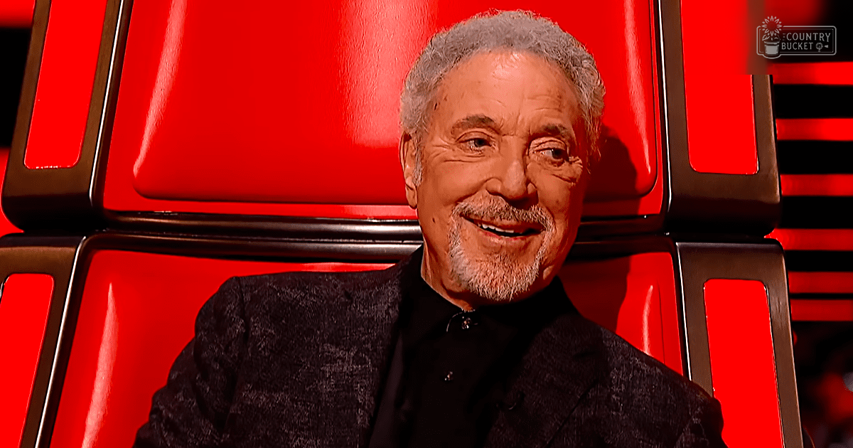 Older adult male in a studio setting, wearing a black textured blazer with a classic mid-20th century style, surrounded by red panels and a metallic chair.