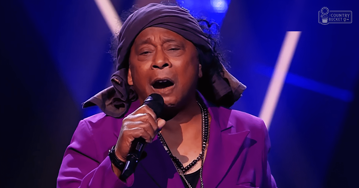 A middle-aged male singer in a purple blazer and black shirt stands on stage, singing into a microphone while wearing a dark beaded necklace and head scarf.