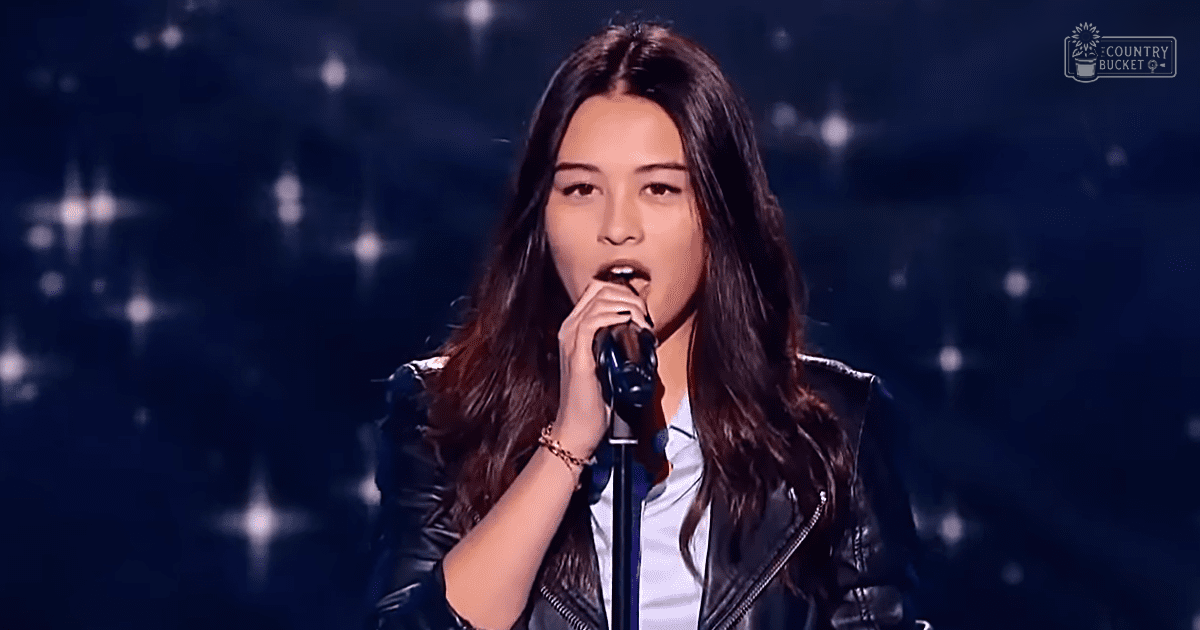 A young adult female in a black leather jacket performs on stage with a microphone, wearing a woven bracelet and an audience visible behind her.