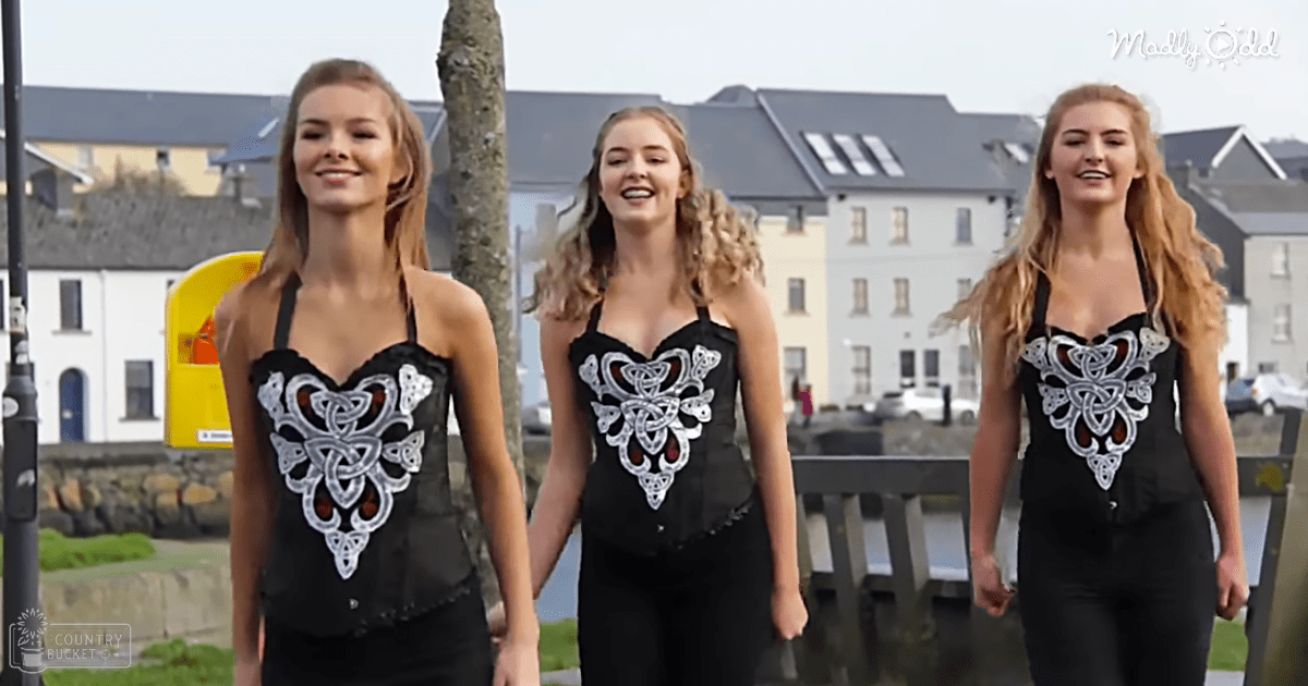 Three young adult women wearing black sleeveless tops with intricate white and silver Celtic knot patterns, standing outdoors amidst suburban buildings and greenery under overcast lighting.