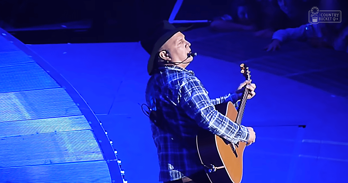 An adult male singer performing on a concert stage with blue lighting and geometric patterns displayed in the background, wearing a plaid shirt and dark cap.