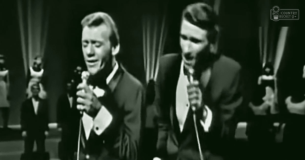 A musical performance scene with adult male performers wearing dark suits, a child holding a microphone stand against stage curtains and lighting backdrop.