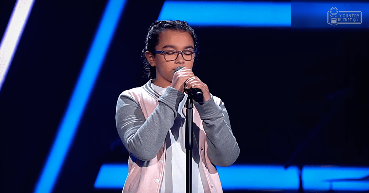 A young child wearing a light pink cardigan with thin black stripes on the collar, performing at an indoor stage setup with blue lighting and modern geometric background.