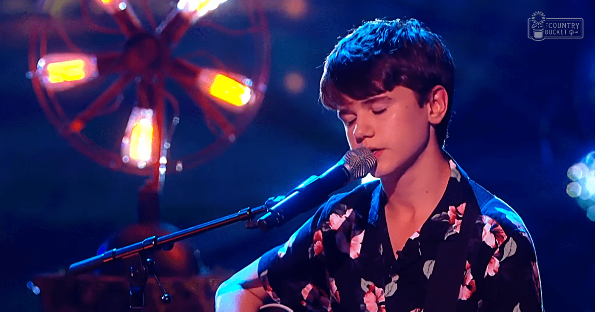 A young adult male singer wearing a dark-colored, floral-patterned shirt with pink, white, and green hues on stage under dramatic lighting.