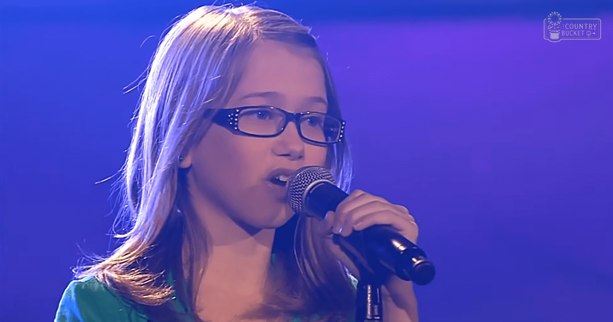 Young adult female singing into a microphone in an indoor performance setting with a green top, blue stage lighting.