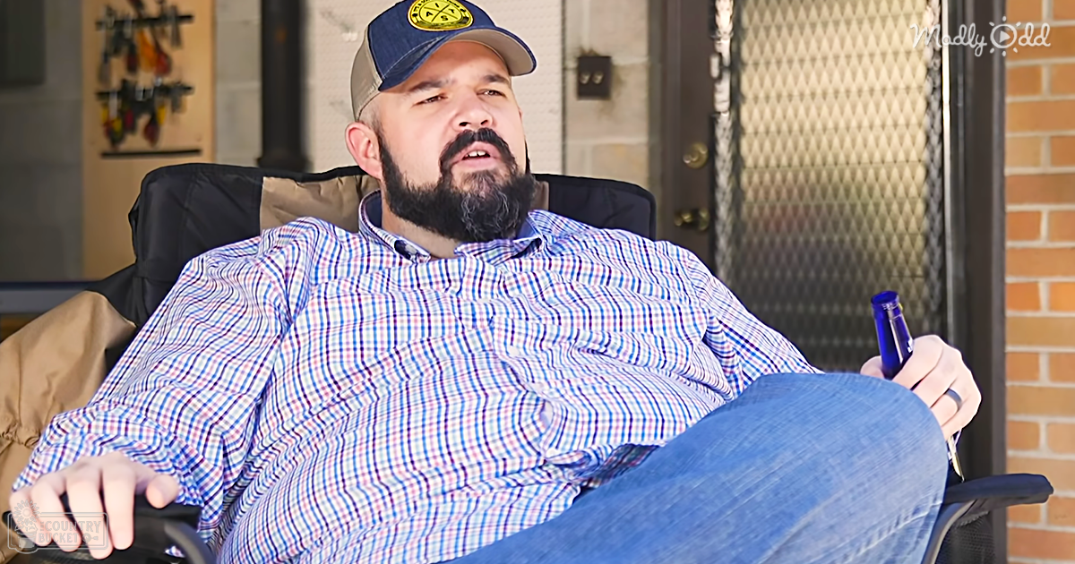 An adult male wearing a relaxed fit blue and white checkered button-up shirt with a collared neckline, holding a blue bottle and wearing a baseball cap in an indoor home or office setting.
