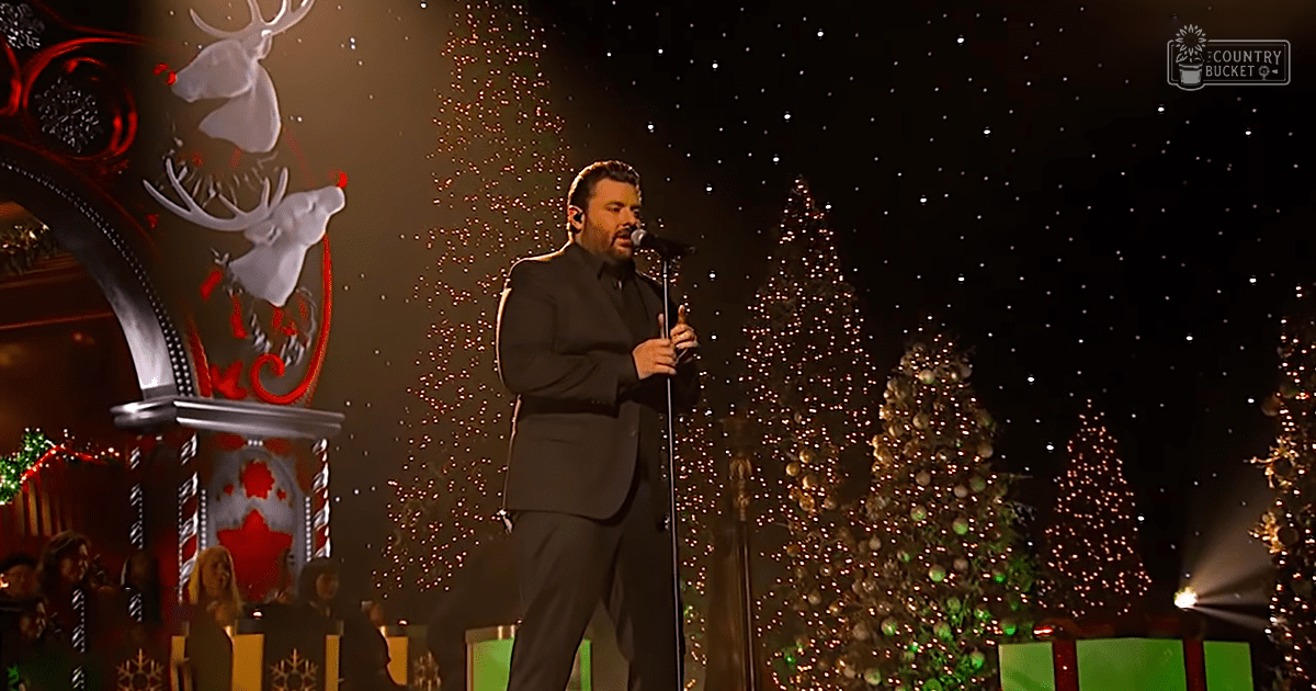 A middle-aged male performer singing at a holiday-themed event with decorated Christmas trees and reindeer illustrations in the background.