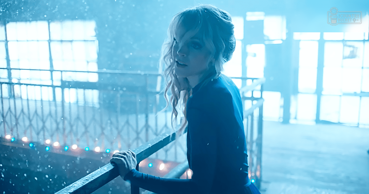 A young adult female in an indoor setting with large windows, wearing a dark blue, form-fitting long-sleeved top or dress with a high-necked collar and smooth fabric. The scene has industrial decor and natural light, suggesting winter.