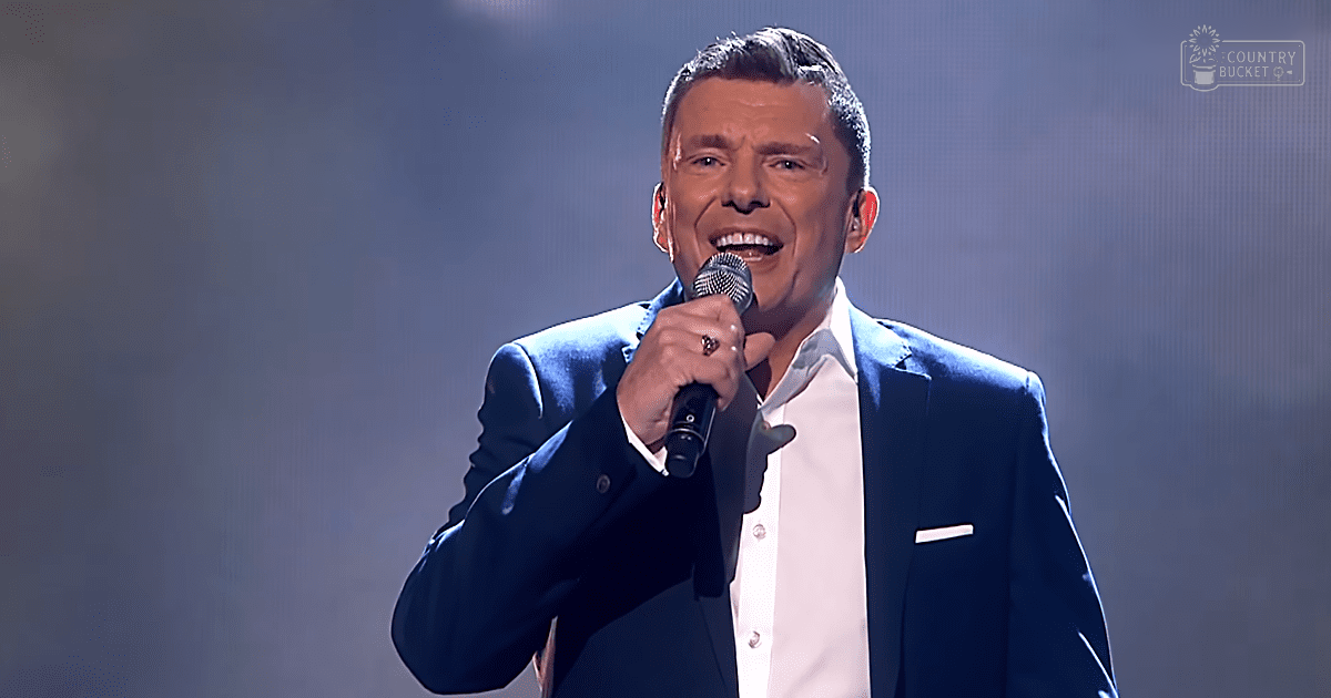 A middle-aged male performer wearing a dark blue suit jacket and white shirt, holding a microphone with 'Madly Odd' text on screen against an indoor stage backdrop.