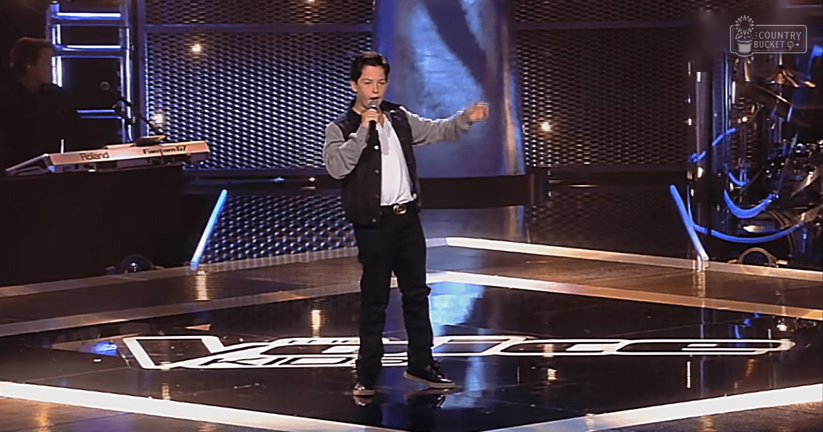 A young adult male singer performing on stage at a talent show with reflective flooring and professional stage lighting.