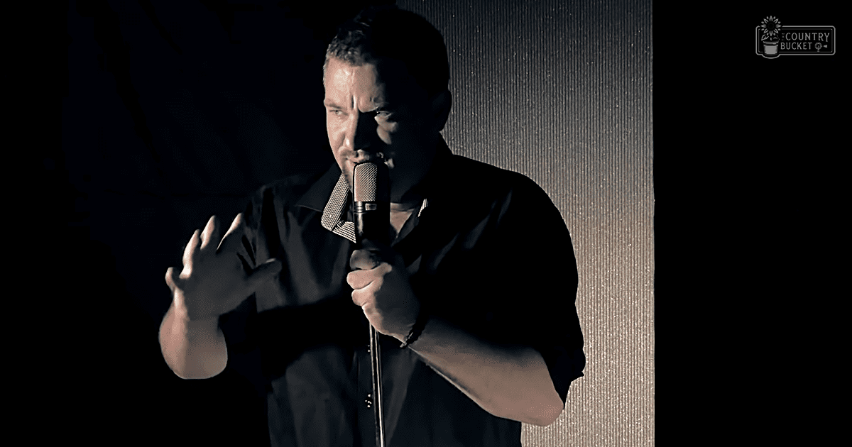 A middle-aged adult male performs stand-up comedy on an indoor stage under focused artificial lighting.