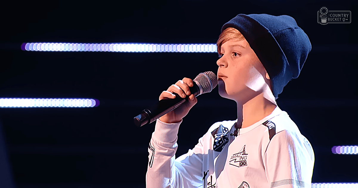 A young child in a long-sleeved shirt with patches and logos, wearing a dark blue beanie hat backwards, performing on a stage illuminated by white and purple lights.