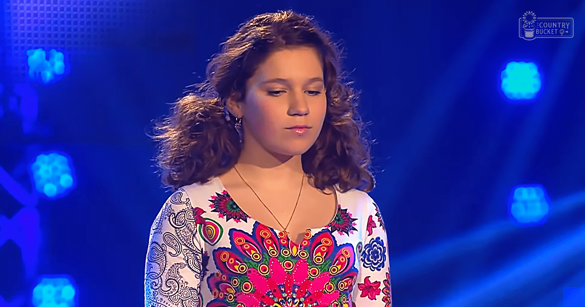 A young adult female wearing a white floral-patterned garment stands in front of stage lighting with 'Madly Odd' text visible. The setting suggests an indoor performance venue.