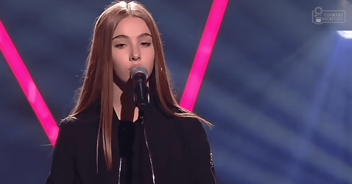 A young adult female in a black zip-up jacket sings into a microphone at an indoor performance stage with bright pink beams and blue background.