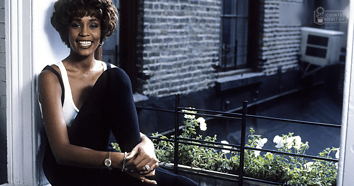 A young adult female wearing a black sleeveless deep V-neck top with form-fitting cut, standing on an urban apartment balcony or patio area during daytime.