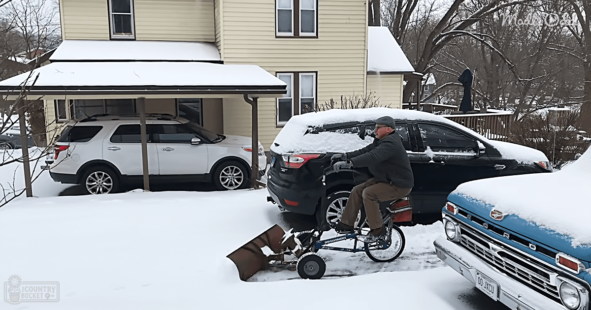 A middle-aged man in a dark gray jacket clears snow from his driveway with a white SUV parked under a carport, surrounded by residential houses and trees in winter, with an overcast light.