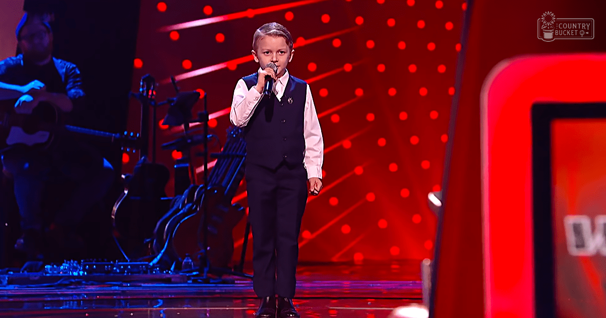 A young male child performing at a talent show stage with red lighting and star-like patterns. The boy is wearing a formal white shirt, black vest, dark trousers, and black shoes.