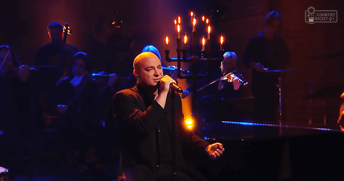 A male adult singer performing on a dimly lit indoor concert stage with a candelabra and piano visible.