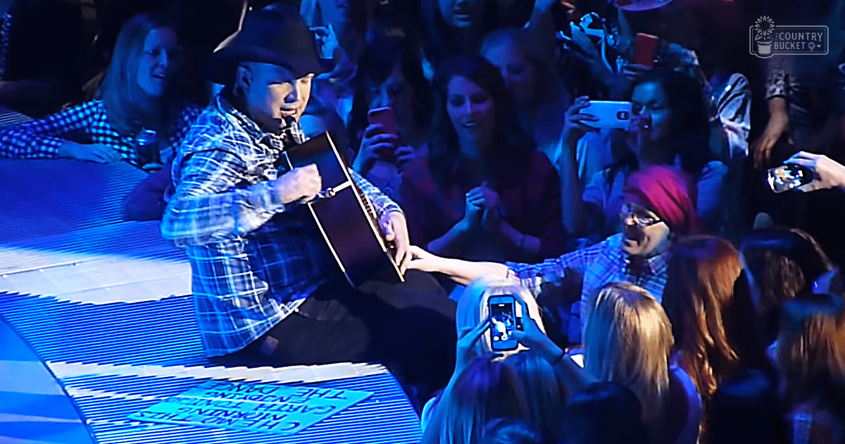 A young adult male singer in a plaid shirt and cowboy hat performs on stage with an engaged crowd holding up phones during a concert at night.