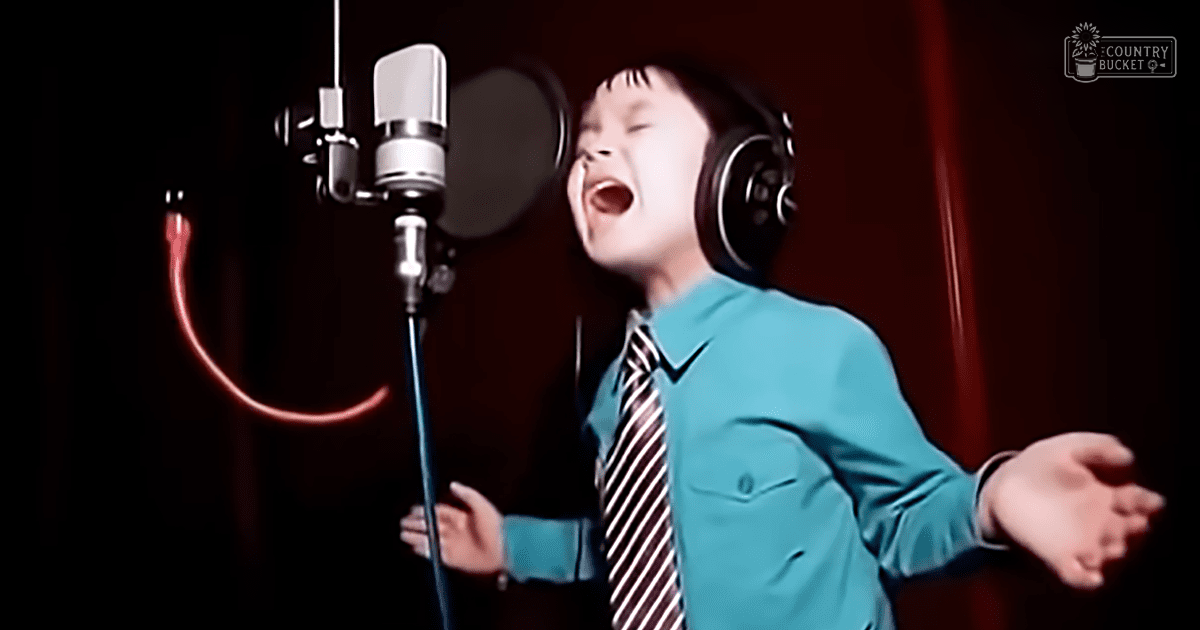 A child wearing a turquoise blue button-up shirt with white stripes on a tie, headphones around their neck, singing in a recording studio setup with red curtains and dark backdrop.