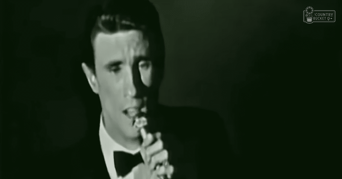 An adult male singer dressed in a formal black tuxedo with white shirt and bow tie, holding a vintage microphone on stage under dim lighting.