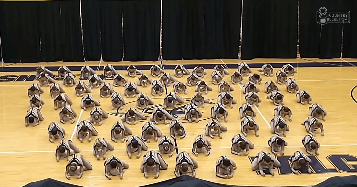Young adults practicing synchronized movements in an indoor gymnasium with a basketball court and black curtains.