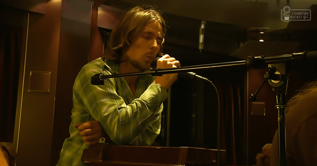 A young adult male singing into a microphone in what appears to be a recording studio, wearing a light green button-up shirt. The setting includes musical equipment and warm indoor lighting.