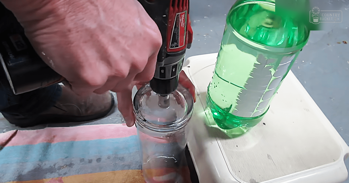An adult male using a drill to modify a glass bottle in what appears to be a workshop or crafting area, surrounded by tools and materials.
