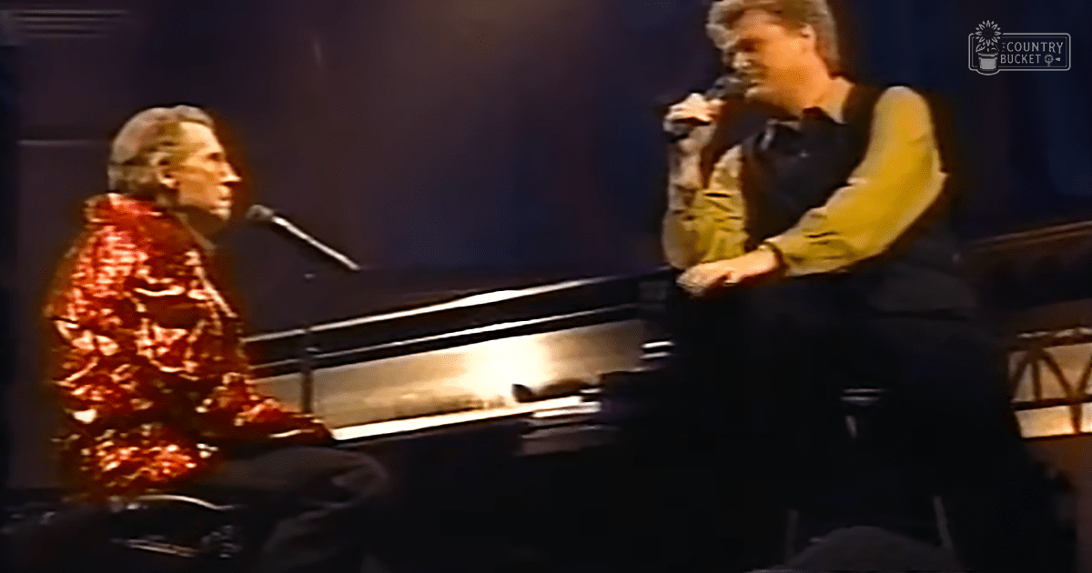 A musical performance in an indoor venue with two male performers; one wearing a shiny sequined floral jacket and another in a muted yellow shirt, set against dark curtains on a stage.