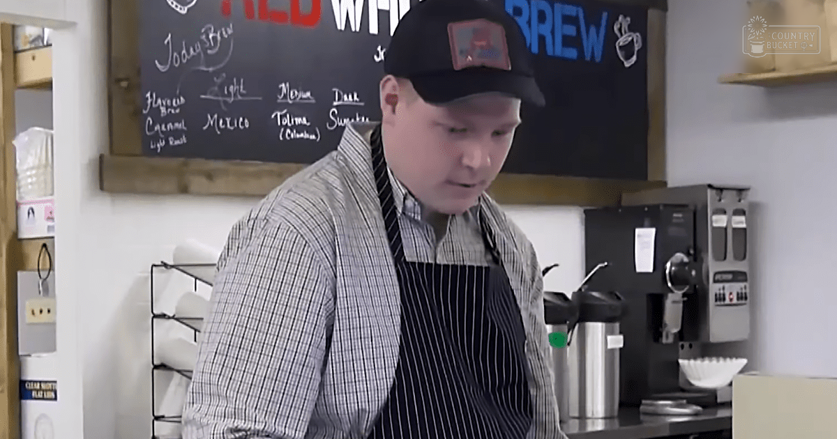 Young adult male wearing a black baseball cap with logo, striped shirt over an apron, working at a coffee shop counter. Blackboard displays brew options and names.