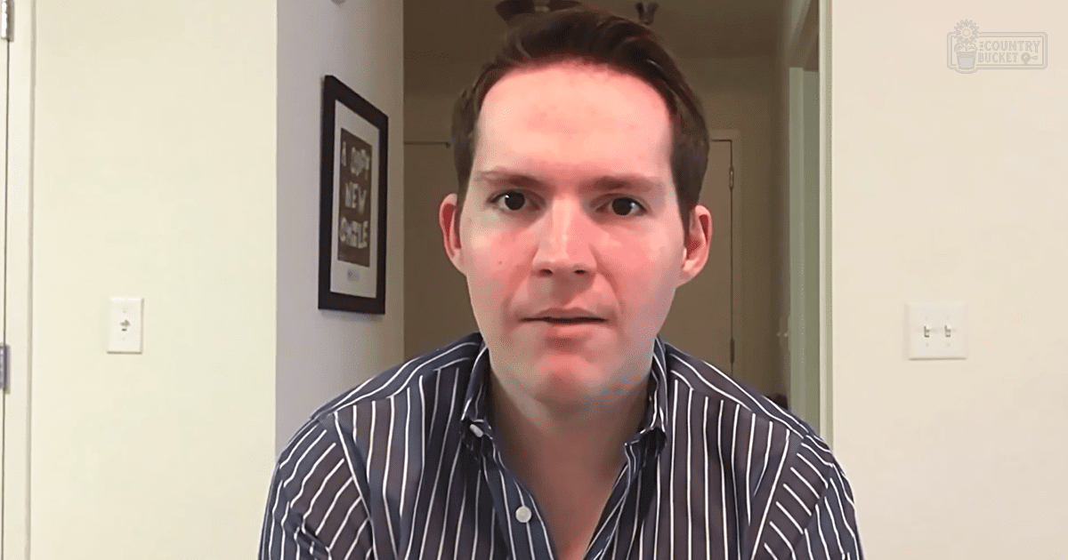 A young adult male wearing a dark blue striped button-up shirt stands indoors against light-colored walls with a framed picture and door frame visible behind him.