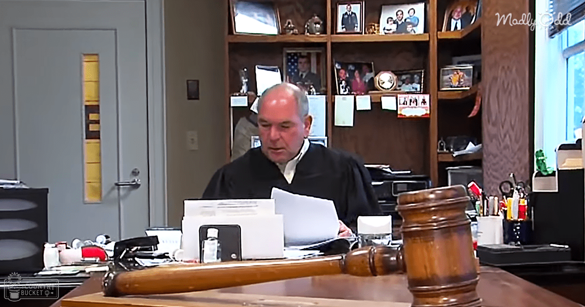 A middle-aged to older adult male judge in a formal courtroom setting with wooden furniture and personal memorabilia on shelves.