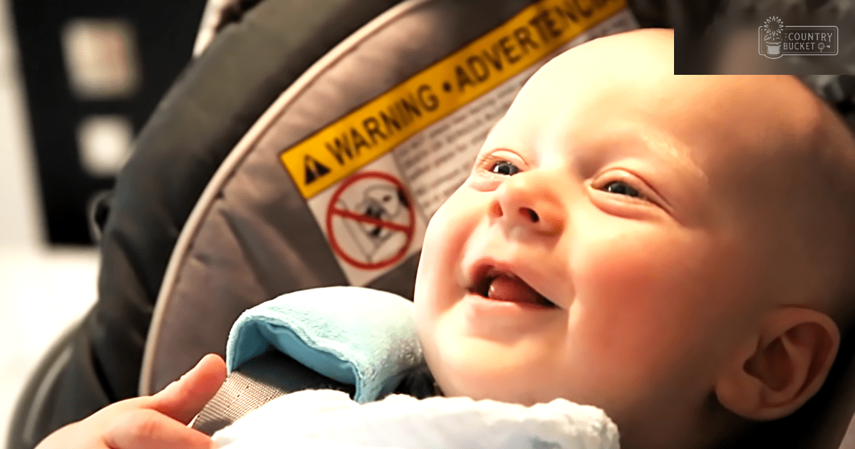 A baby in a car seat, wearing a light blue bib or blanket indoors with warm natural lighting suggesting daytime.