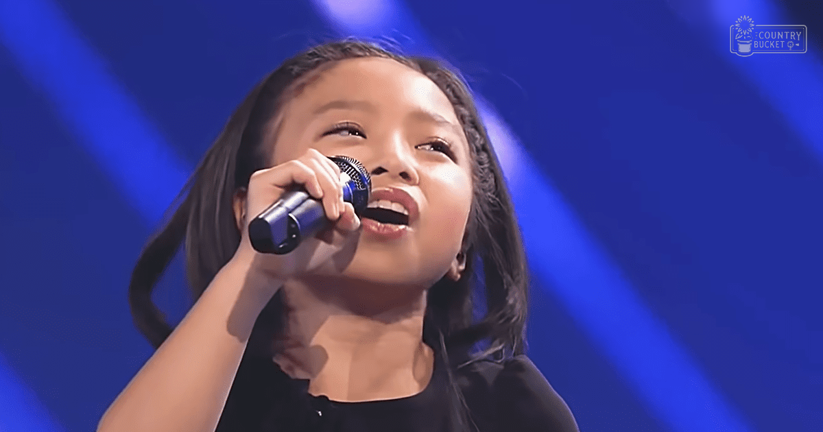 A young female child sings into a blue microphone on a stage with professional lighting, wearing a black sleeveless top or dress.