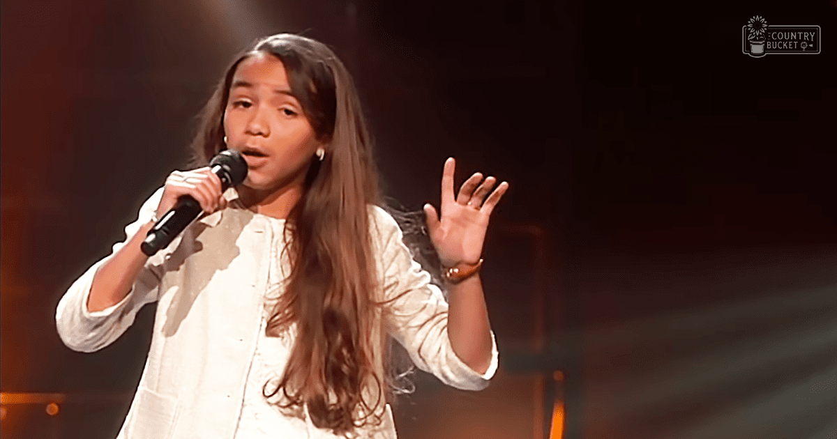 Young adult female singer wearing a loose, white collared blouse with long sleeves rolled up slightly, holding a microphone on stage under dim performance lighting.