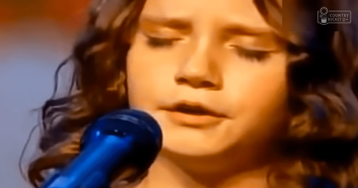 A young adult female singer wearing a light beige blouse performs into a blue microphone on stage at an indoor evening event.