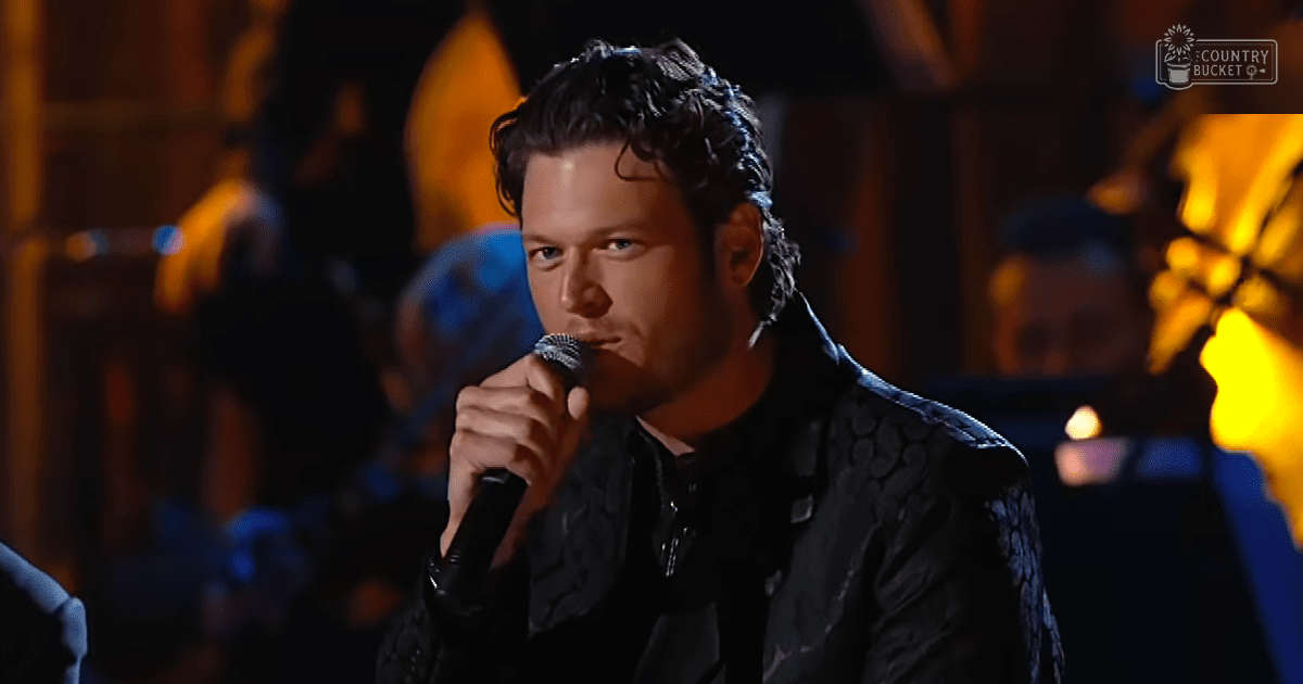 An adult male singer wearing a textured dark jacket with long sleeves and high collar, holding a microphone on stage under warm, dim lighting.