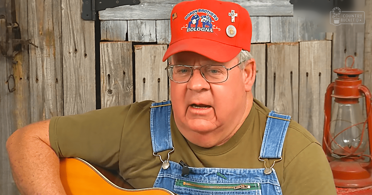 An older adult male in a rural setting wearing a red baseball cap with 'Morov Brothers Bologna' text and logos, blue overalls, and glasses. A lantern is present on the right side of the frame against weathered wooden planks.