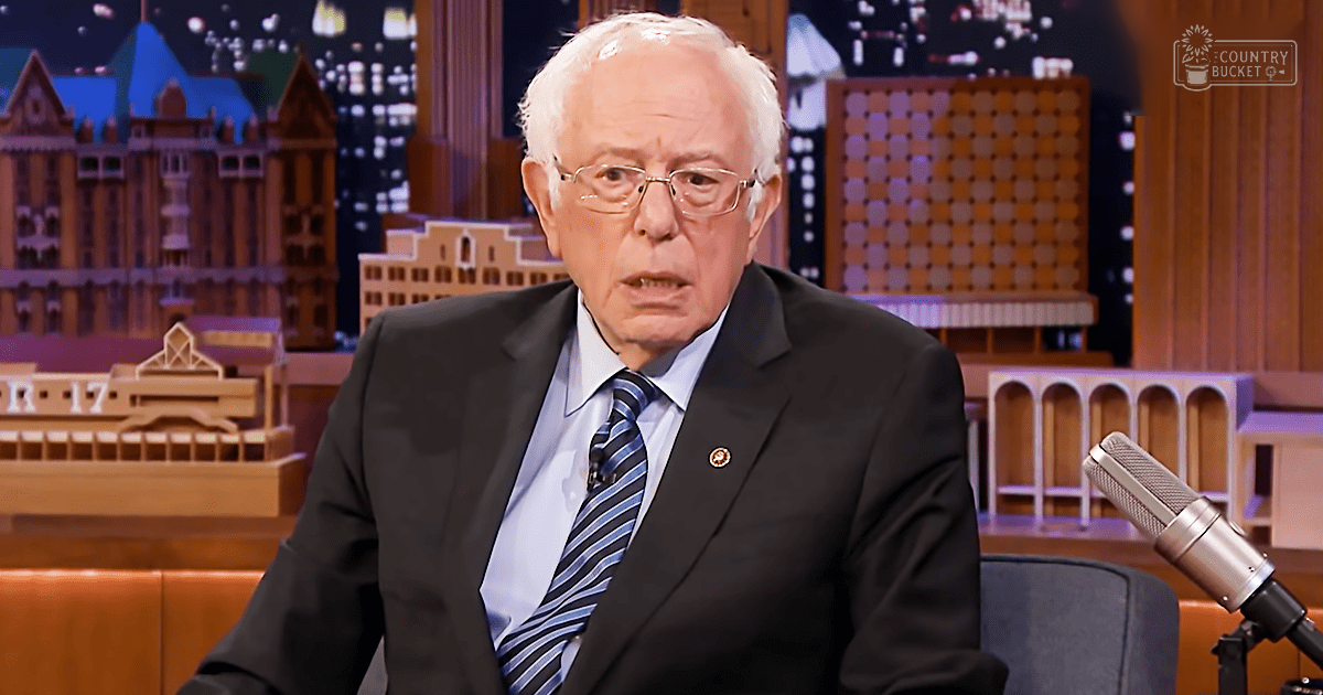 An older adult male wearing a dark suit jacket with a lapel pin, light blue dress shirt, and striped tie in a studio setting with cityscape backdrop at night.