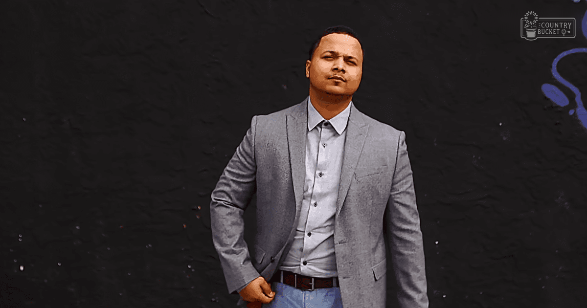 A young adult male in a light gray blazer and white button-up shirt stands against a dark wall with faint blue graffiti-like markings, suggesting an urban setting during the daytime.