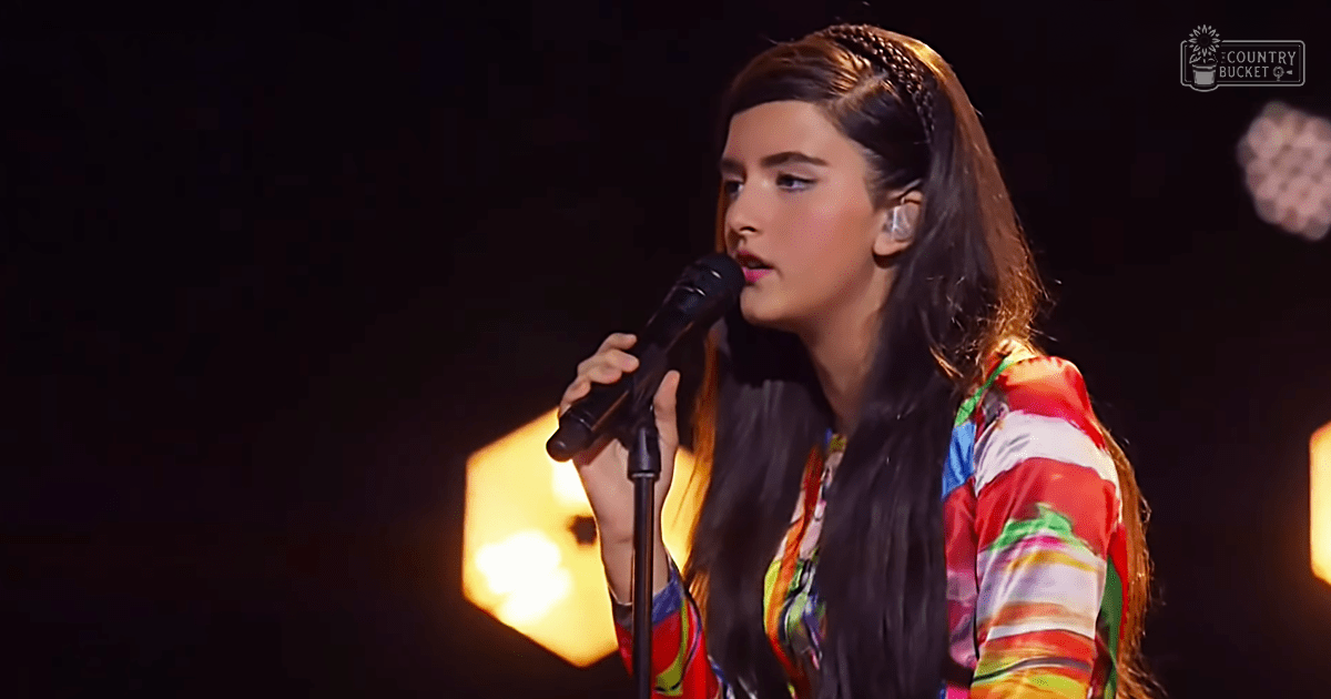 A young adult female sings into a microphone at an indoor performance setting under stage lighting.