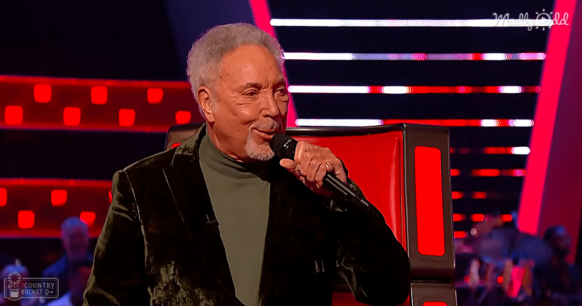 A middle-aged man in a dark textured jacket and green turtleneck stands on what appears to be a stage with red and white illuminated lines, suggesting performance lighting.