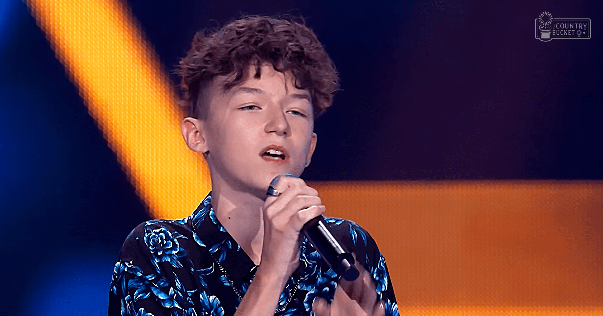 Young adult male singer wearing a dark blue floral shirt with bold patterns, holding a microphone on an indoor stage illuminated by bright orange lights.