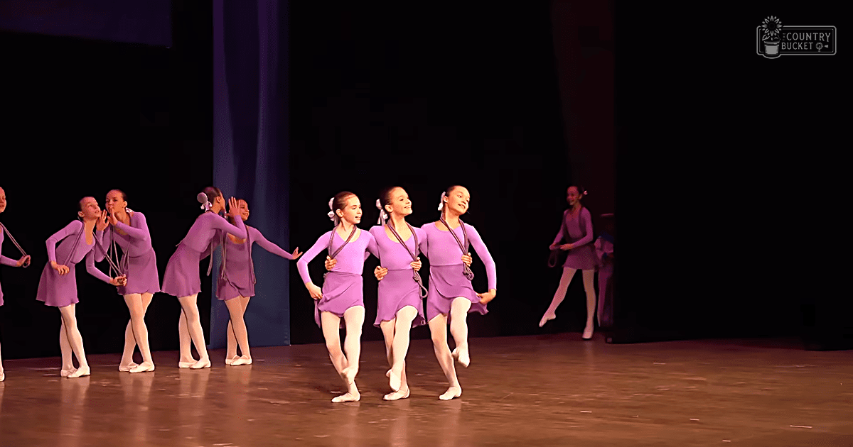 A group of young female dancers in form-fitting, sleeveless purple leotards with attached skirts perform on a dimly lit indoor stage with dark curtains and wooden flooring.