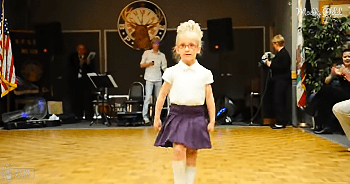 A young girl in a school uniform walks through an indoor event space with adults and musical equipment visible. The setting suggests a community or school function.