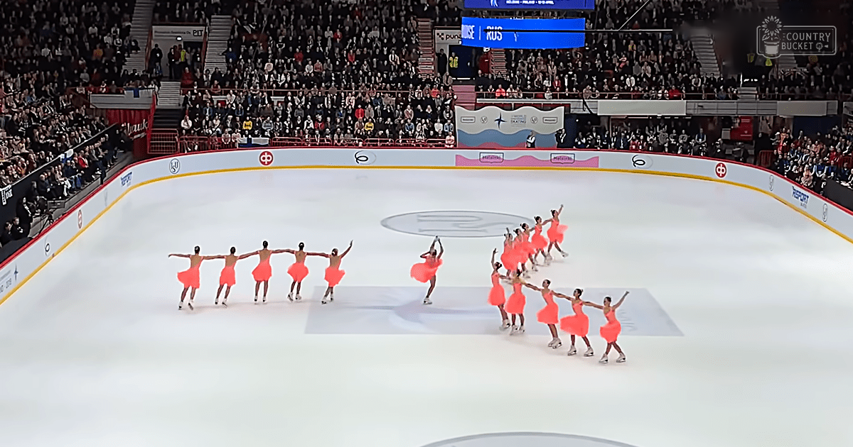 A synchronized skating team in bright orange dresses performs at an indoor ice rink with a crowd of spectators seated around it.
