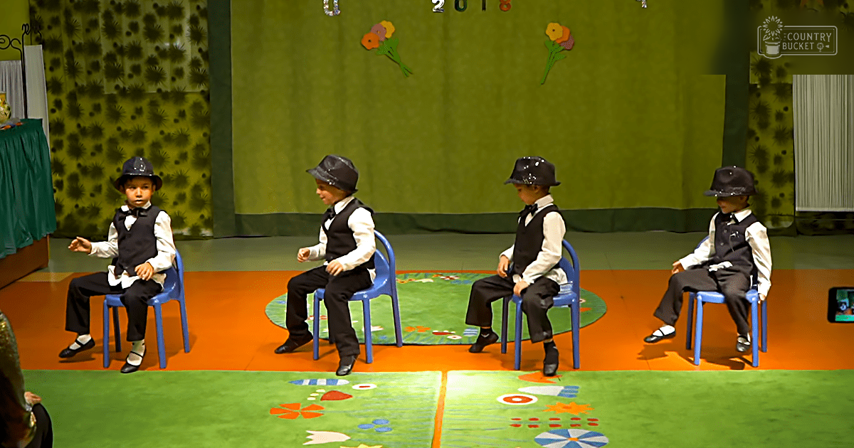 Children in a school setting, performing with formal attire on a stage decorated for spring or summer.