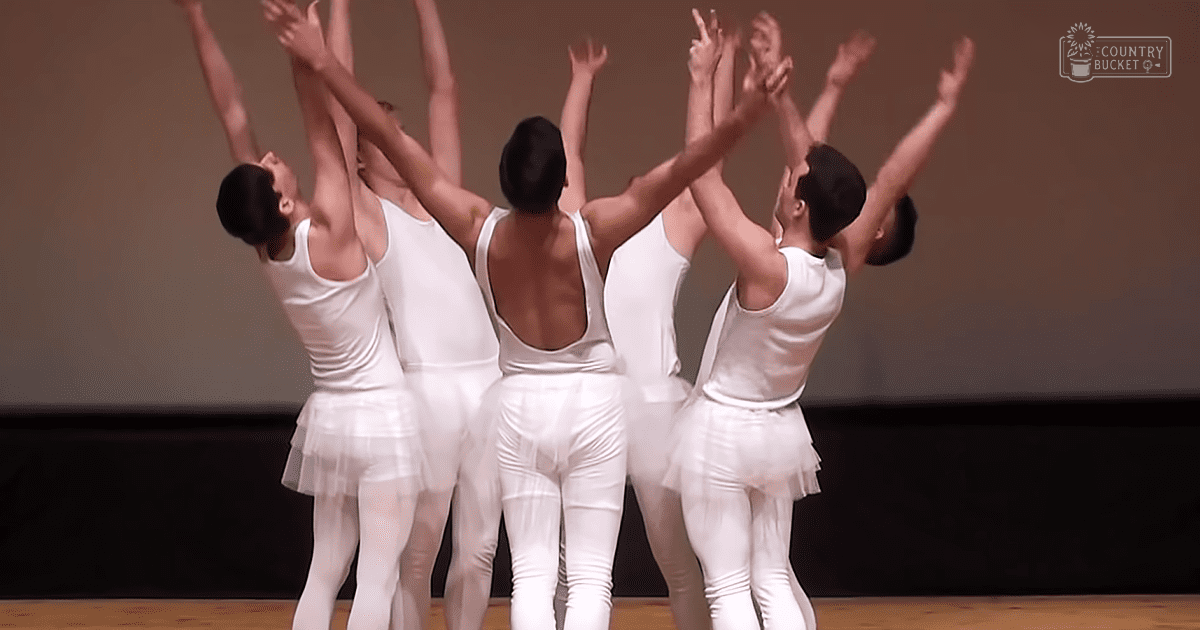 Young adult female dancers in white leotards with cutouts performing on a simple stage under indoor lighting.