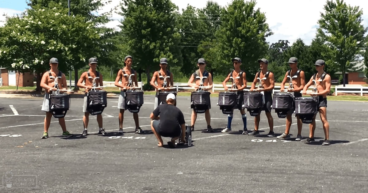 A group of young adult males practicing with drums in an outdoor parking lot setting during the day under natural light.